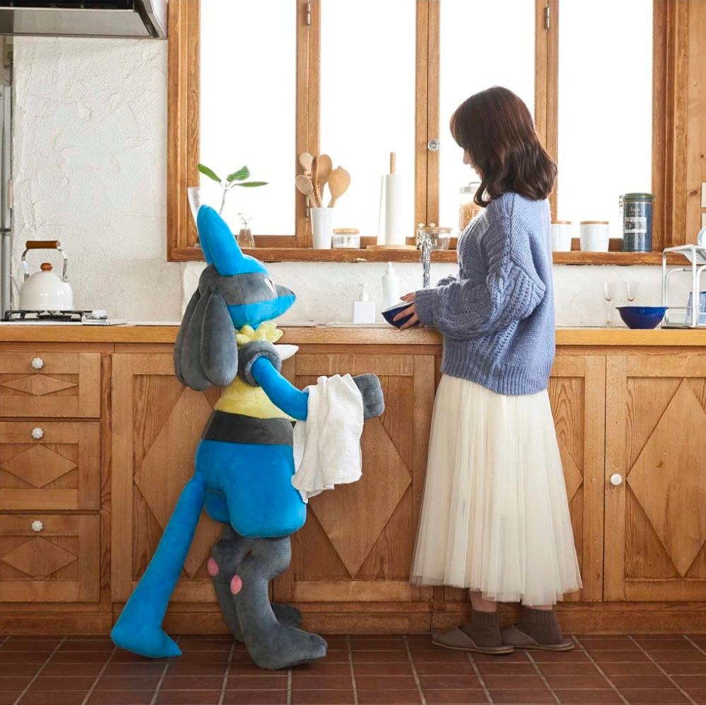 This is an image for a Pokédex size Lucario from the Pokémon company in the picture. The 4 foot tall plushy is holding out a dish towel for a woman who appears to be doing the dishes.