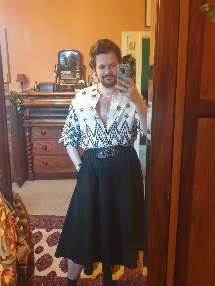 A white man with short brown hair taking a selfie in a mirror. He is wearing pearl necklance and a half-open white shirt with black embroidery, tucked into a calf-length black skirt. 
