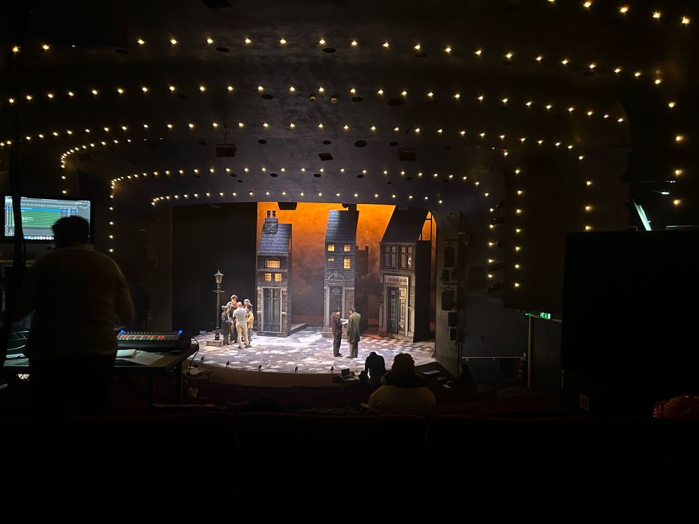 The stage at derby theatre - an orange sky with three tall thin houses looming over the cast who are stood around the stage waiting for tech to start 