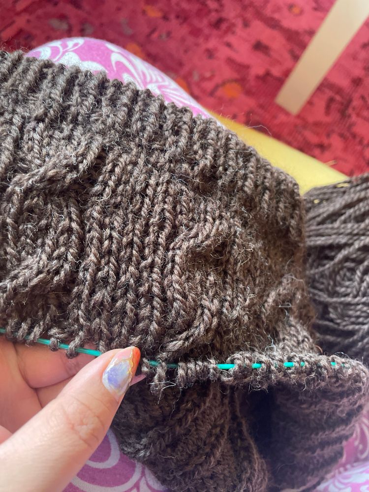 A tan hand with short nails and nail blue and red nail art holds a short section of dark brown unfinished knitting. Ribbing is visible at the top and a teal circular knitting needle cable is visible peeking through the live stitches at the bottom. The knitting pattern a 4 by 1 ribbing pattern with long vertical sections without cables alternating with sections that contain single twist cables.