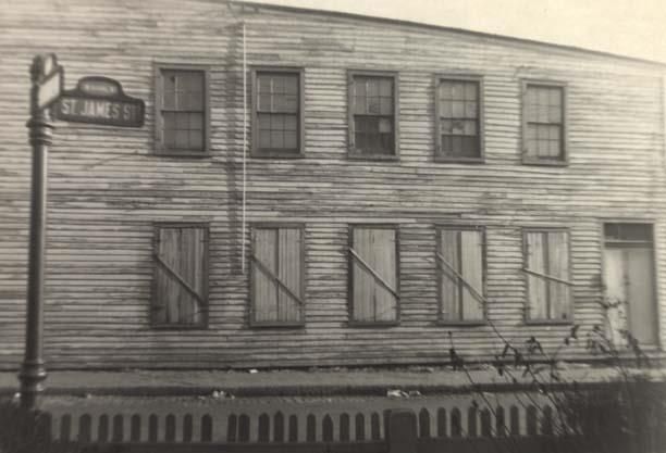 Black and white photograph of side of two-story wood-framed building with lower level windows barred; street sign on sidewalk at left; street and picket fence in foreground; "at West Duval" written at bottom of photo; "93-4 / Feb. 1953 / St. James St. at W. Duval" written on reverse; Richmond, Virginia.