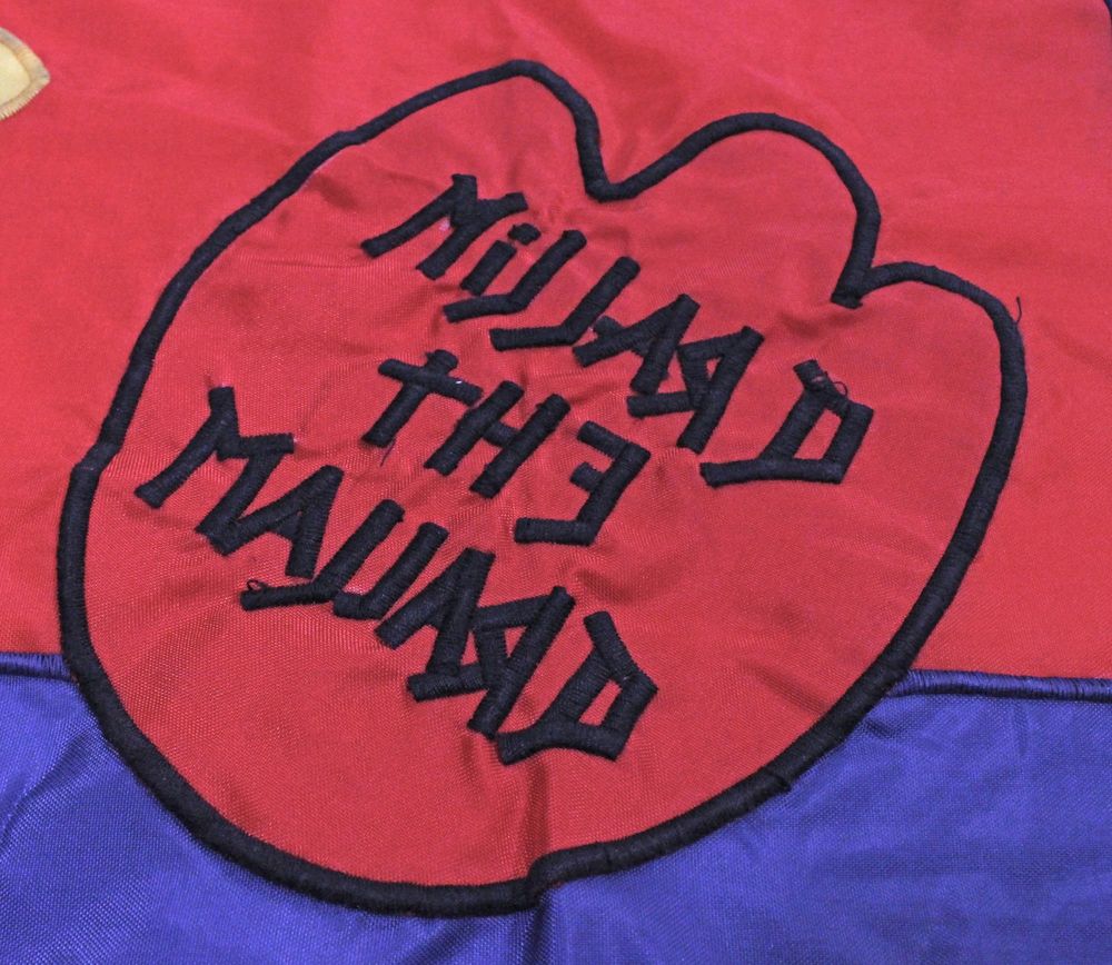 Flag of synthetic fabric in red with a blue border, appliqued with a duck mascot in yellow, green, and brown, with leather tabs for hanging as a banner: rectangular; pole sleeves at top edge; reinforced hanging tabs inside sleeve at R and L edges; figure at center; logo at bottom L corner; 2" border; hemmed perimeter.