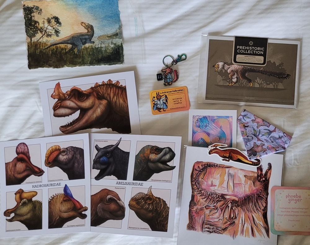 watercolour of a therapod looking to the rising sun, prints of ceratosaurus, various hadrosauridae (my babies), various abelisauridae, utahraptor, small pin of a raptor geneticist wearing a cute lab coat, carnotaur sticker, print of a tyrannosaurus skeleton, and a pet bandana covered in mini dinosaurs