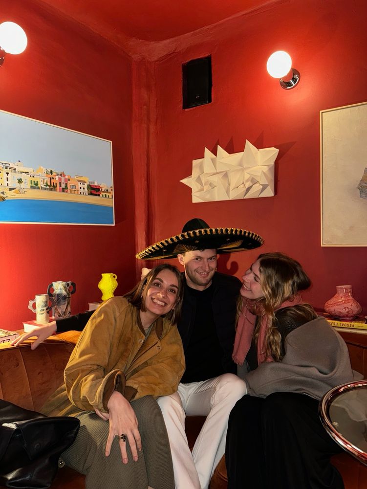 Drei Personen auf einem Retro-Sofa vor roter Wand – ein Mann mit großem Hut, flankiert von zwei Frauen. Stilvoll und zeitlos.