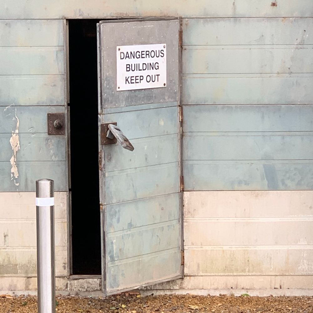 A half open door of an industrial building. A sign on the door reads ‘dangerous building keep out’ 