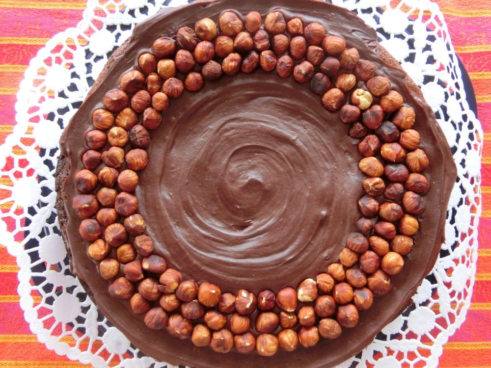 Blick von oben auf eine runde Torte mit Schokoganache und drei Reihen Haselnüssen kreisförmig aufgelegt. Weiße Tortenspitze.