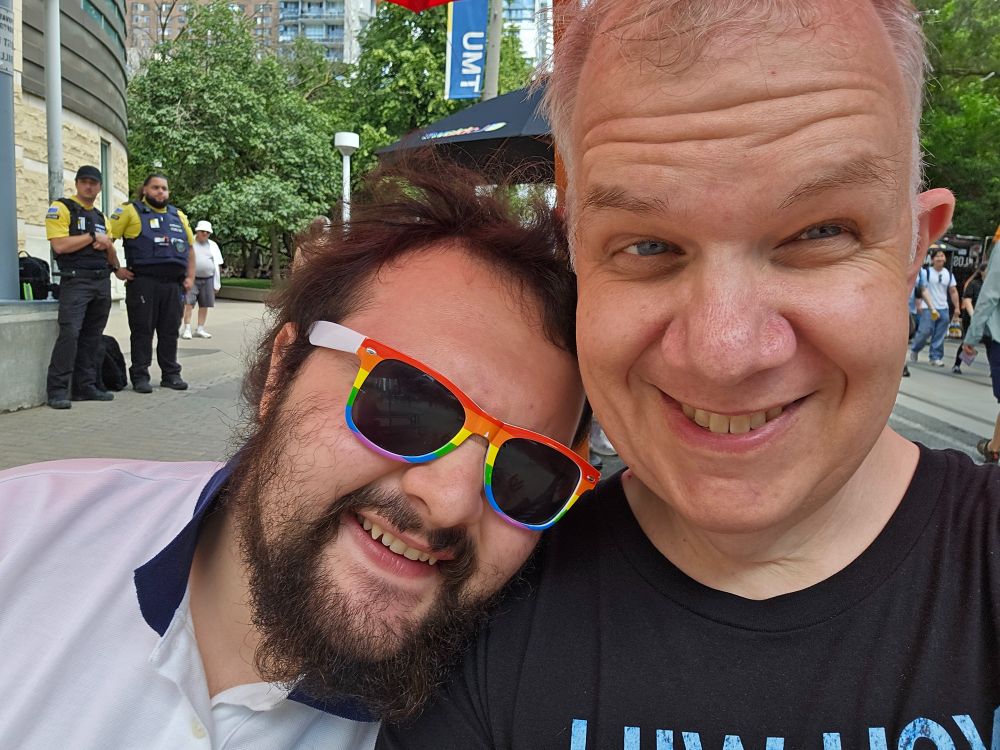 The poster, a middle-aged white man,  sits with his son, a young white man with a beard, wearing and rainbow sunglasses, at a bench at Toronto Pride 