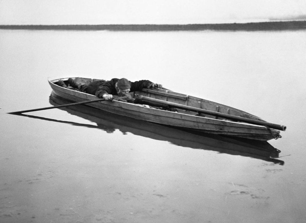 Sighting a ‘Punt Gun’.  Black and white photo of a very large shotgun (>6 ft long, ~2 in. diameter) in a small rowboat.  A man is laying down in the boat behind the gun aiming the gun by moving the boat.