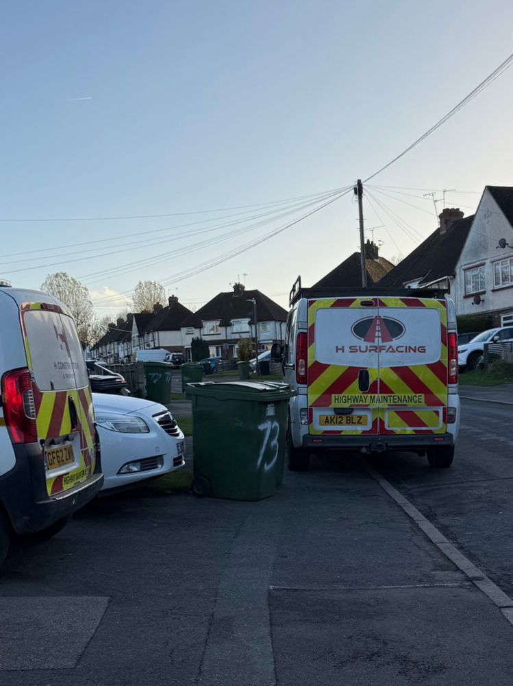 A lorry parked off on the pavement, next to 2 wheelie bins next to a car with its front sticking out from the driveway onto the pavement. The pavement is completely blocked. 