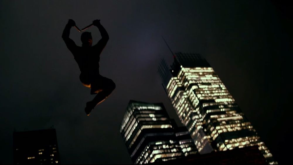 still from Daredevil television show. shot is pointing up at a night sky with lit up sky scrapers jabbing in from the bottom right and daredevil, in red and black silhouette leaping down from above the camera billy clubs raised above his head. it's very noire and the shot is lifted right from the comic books.