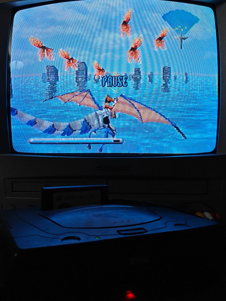 A Sega Saturn in a small table in front of a CRT television. On the screen of the TV is the first level of Panzer Dragoon 