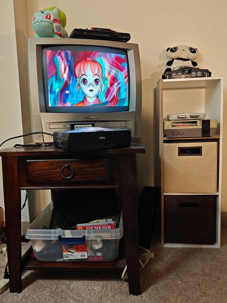 A CRT TV with a built in VHS DVD player is on top of a small table with a Sega Saturn in front of it and a Sega Genesis on top. In a bin on the shelf under the table are my most frequently played NES, Genesis, SNES, and N64 games and a DVD binder