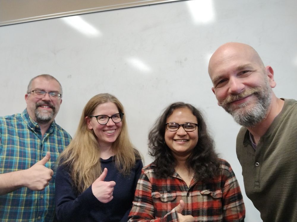 From left to right: Dr. Dave Westerman, Dr. Lisa Arnold, MA graduate Mahmuda Rahman, and Dr. Zoltan Majdik, all smiling and giving a thumbs up, standing in front of a white board.