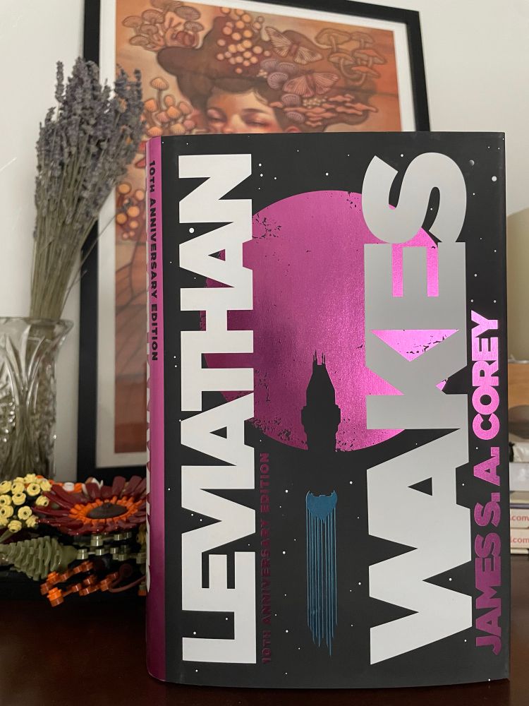 The 10 year anniversary edition of Leviathan Wakes by James S. A. Corey. On the cover the title is vertically displayed in white block letters. A rocket blasts off in front of a pink celestial sphere. Partially behind the book is a framed art piece and Lego flowers. Off to the side there is a dried lavender bunch in a glass vase. 