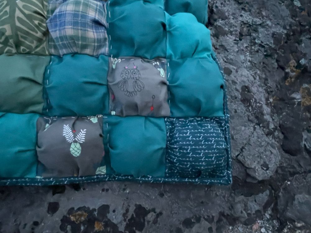 Corner of a puff quilt on a dark grey rock at the beach. The puffs are teal green colours and the bottom right one is teal with a white script saying fuck cancer 