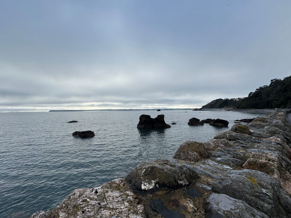 In the foreground a deep sea wall covered in lichen, then the sea with some large rocks is still and greeny blue. The top of the photo shows heavy grey and white clouds with a sliver of light along the horizon.