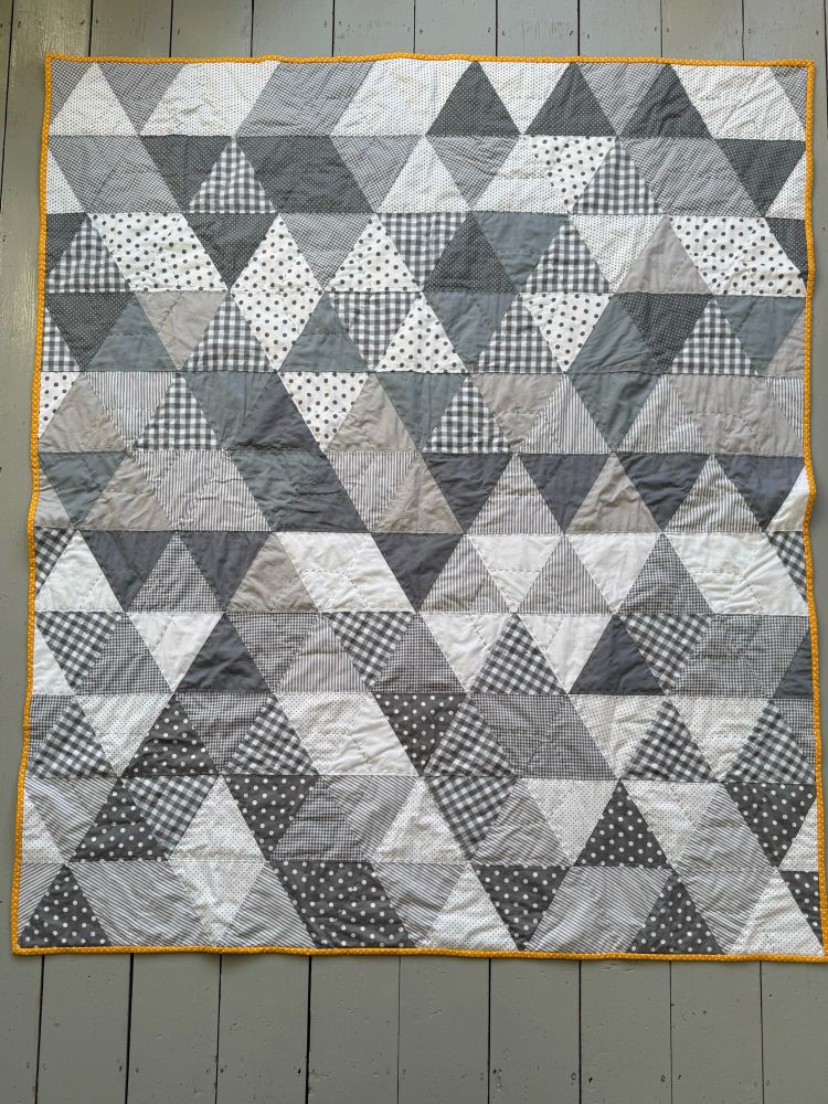A large home made quilt made up of monochrome grey and white fabrics cut into triangles randomly placed. It has a bright yellow polka dot binding around the outside. 