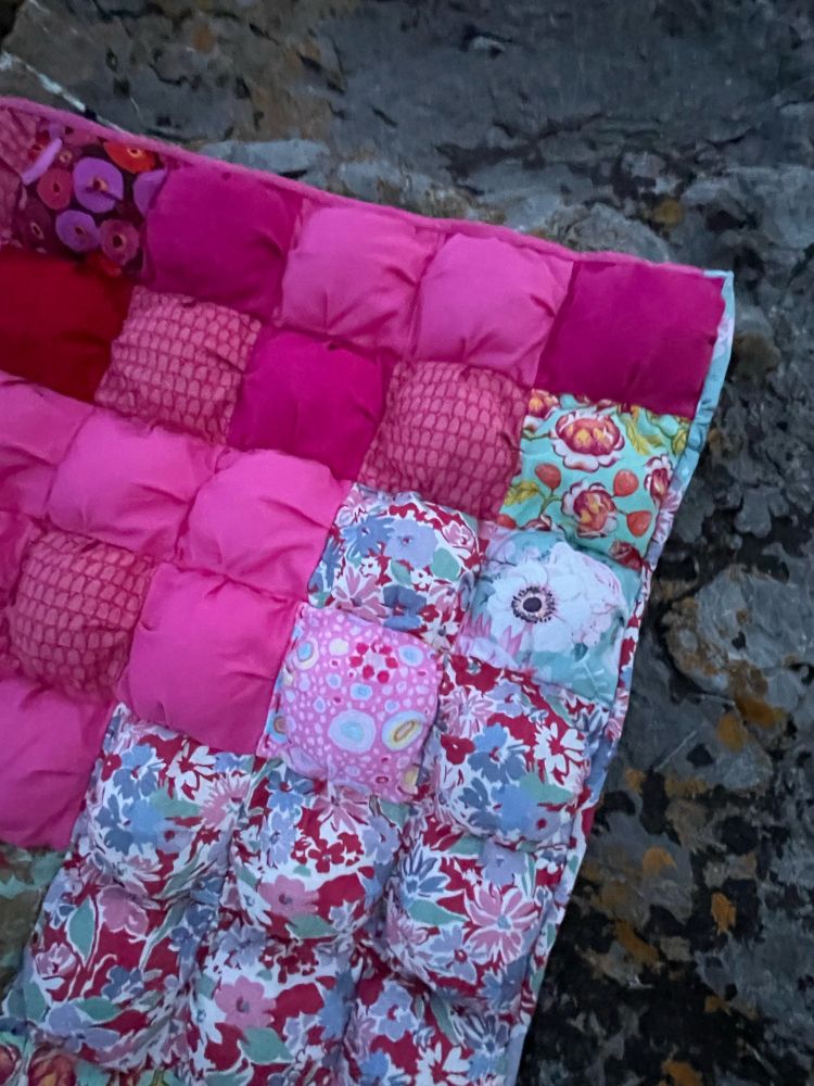A corner of a puff quilt with puffs made from various pink fabrics and floral fabrics. It is sitting on a dark grey rock at the beach 