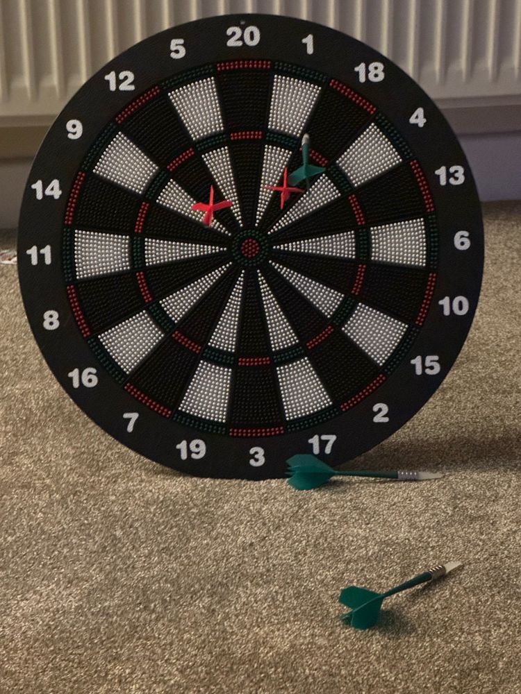A child’s safe dartboard has one green and two red darts in it and too green darts on the floor. It’s on a beige carpet, in front of a white radiator