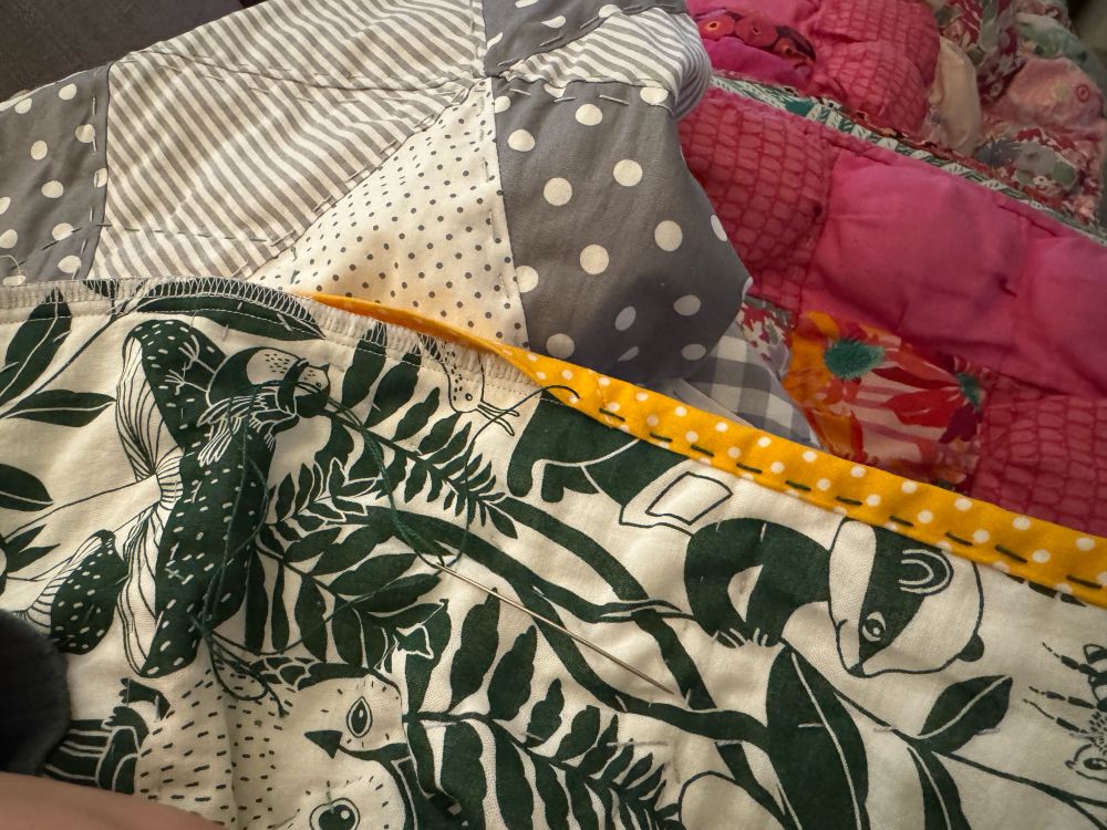 A green and white quilt backing with a yellow polka dot bonding being hand sewn on. Behind is the front of the quilt made of grey and white large triangles. Underneath that quilt is a puff quilt with pink puffs on show.