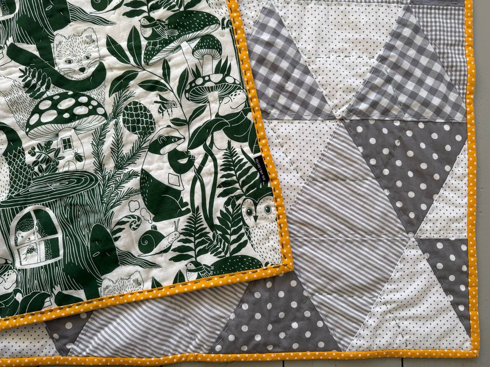Close up of the front of a quilt with the back turned over. The front shows hand quilting in rows where the grey and white triangles are sewn together and in hexagons. The yellow binding with white polkadots pops.