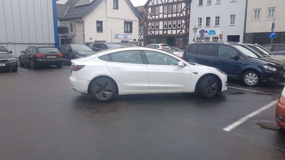 Parkplatz. weißer Tesla steht diagonal auf Parkplatz. Im Hintergrund andere Autos und Fachwerkhäuser