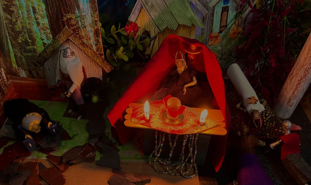 Exterior, night, woods. In the background are a few tiny looking houses with A-frame roofs, some trees with eerie white light or smoke in them and some dark vegetation. 
On the right, between a whitish tree trunk and a red tent, is a bier with a girl laying on it. It’s ALS as the Dead Girl, wearing a long white dress with a yellow flower on her chest, her head towards us, lying on a 
blanket of red, gold and brown patterns. In front of her bier are 3B and Raven as Mourning Women, knelt down before her. 3B in a brown shirt and purple skirt, Raven in a red shirt and gray skirt.
Left of this tableau, in the middle of the shot, is a red tent behind FLO as Old Woman. She’s in flowing black. On the table in front of her is a piece of red and green heavy fabric. On the fabric is a circle of some white powder surrounding an orb in a metal goblet, nestled in red cloth. On either side a small lit candle, creating a small area of light in the darkness.
On the far left is the end of the scene. In a darkness slightly lit by the candles in the middle, Ralph G. as a Man stands looking down at R2-B1 as newly ensouled Angel. Ralph G. is in purple pants and a white 
floofy shirt with a brown sash. Angel is in all black and prostrate on the ground.