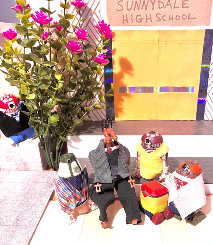 Exterior, day. Sunnydale HS front steps. Bright lighting. The doors are closed. A bush with pink flowers is on the left. Behind the bush on the left is R3-T2 as Buffy, in blue pants under a black jacket. 
On the school steps are R4-M9 as Xander, IRRI as Giles, R4-P17 as Cordelia, Bee as Willow and R4-H5 as Oz. Xander is in a blue and red plaid shirt. Giles is in a gray suit jacket and black vest and pants. Cordelia is in a bright yellow dress. Willow is in lime and orange. Oz is in a white tee with a red graphic under a pink shirt. 
They are gathered close together. No one else around. They don’t see Buffy. Behind the bush, Buffy looks at them but doesn’t approach. 