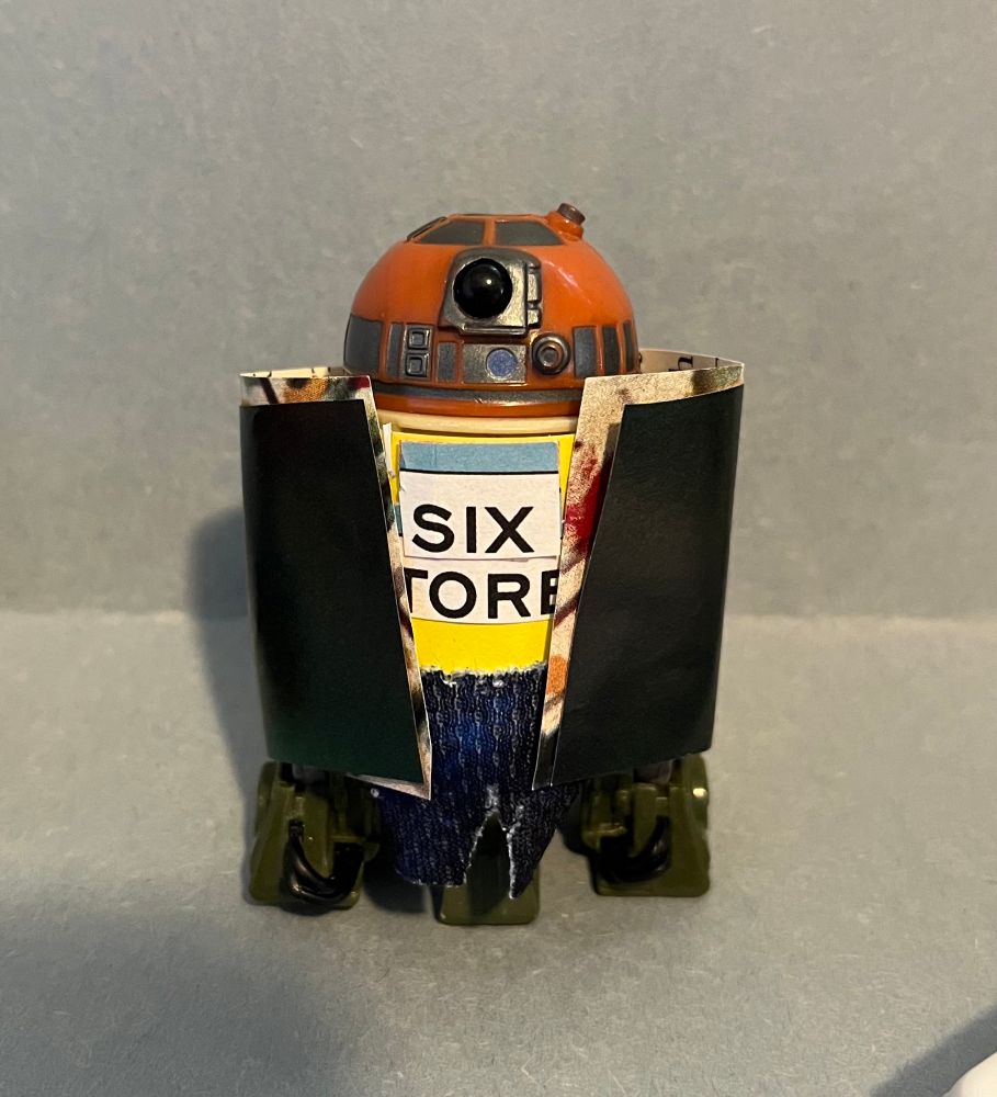 Here Oz has a paper yellow tee with a blue stripe on top of black typed text on white paper, “six tore” all we can see. Cloth blue bottoms. A paper multicolored shirt mostly covered by a paper coat of dark green that looks like a dirty black. 