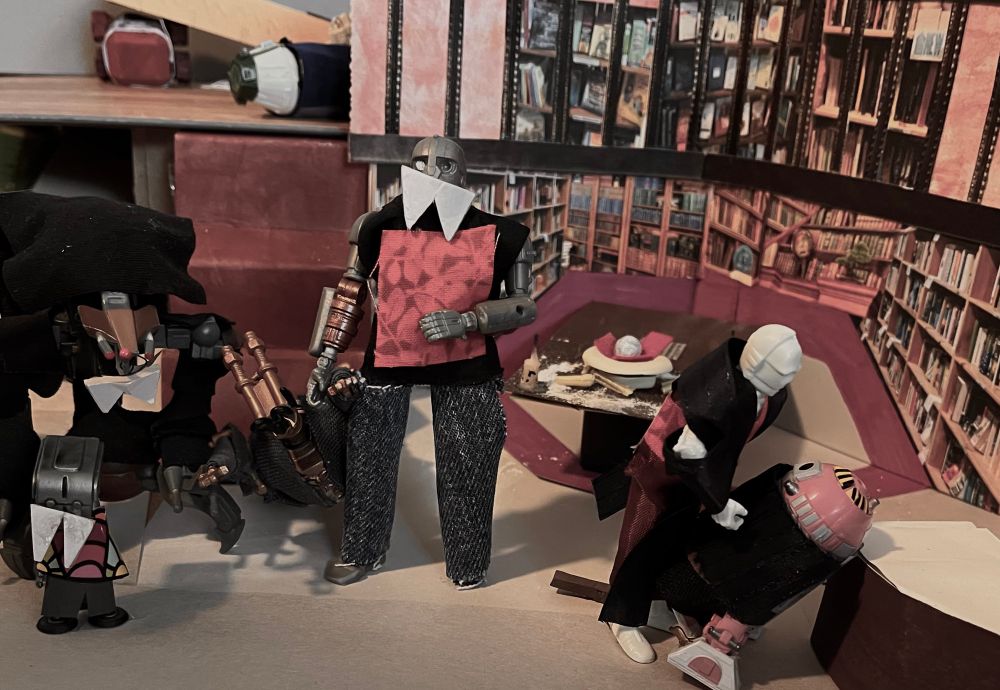 D: Interior, Sunnydale HS Library. On the left up the stairs we can just see Bee as Willow, unmoving under the fallen bookcase, and R4-M9 as Xander lying still at the top of the stairs. 
Below them, Vampires Remy, TODO, and SM-33 are dragging IRRI as Giles out. 
On the right, K-3PO as Drusilla is bending menacingly over Kit as Kendra, who is no longer fighting, just staring up at Dru.