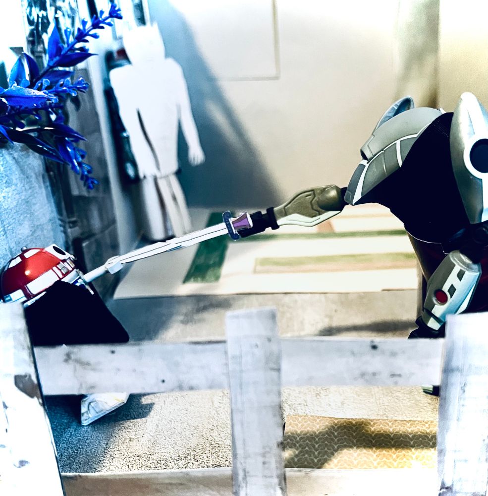 B: Exterior, Angelus’ Mansion balcony. In the background we can just make out the room inside with Acathla as a blurry figure. On the left is a short wall with a purple bush growing over it. In the front of shot is a short railing.
Against the low wall is R3-T2 as Buffy. Her sword is gone. She’s partially collapsed back against the wall. Standing in front of her is Blu as Angelus. He still has his sword. He’s lunged it right at Buffy’s face. But she has somehow extended one small manipulator at the exact time to catch and stop the sword. 