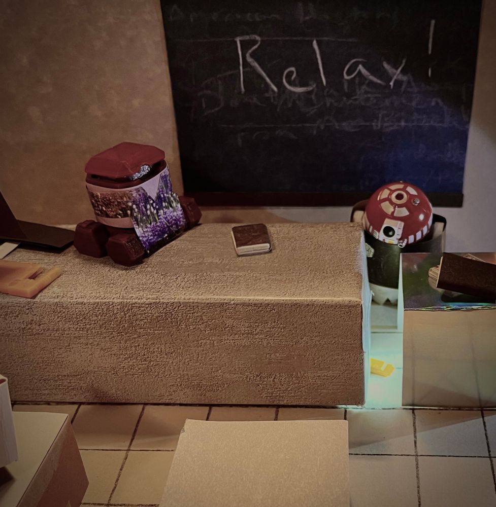 Interior, computer classroom, somewhat dimly lit in afterschool hours. On the gray back wall is a chalkboard with the word “Relax!” written on it. A gray teacher’s desk is in front of that. On the far left we 
can see part of a black keyboard and a yellow piece of equipment, a drive maybe. Bee as Willow is on the desk, next to a book, in a lilac shirt under a smock of purple and white flowers. She’s looking to the right 
at R3-T2 as Buffy, in a black shirt and white pants. Buffy’s staring down at a yellow disc seeming almost to exude light from where it sits on the floor between the desk and a silvery file cabinet next to it. 