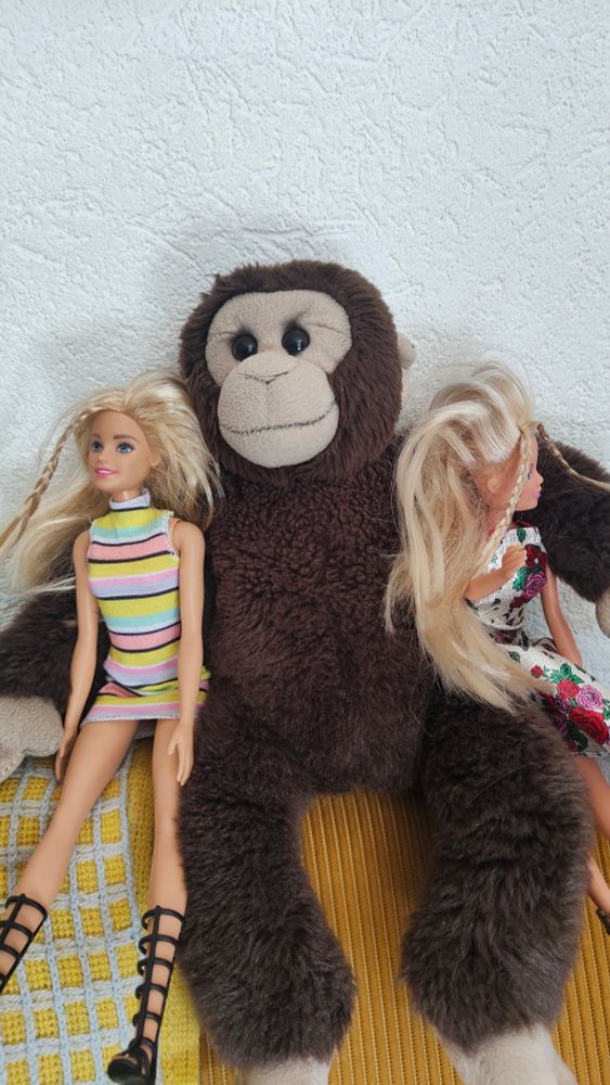 Ein stoffaffe hält zwei  blonde Barbiepuppen im Arm.