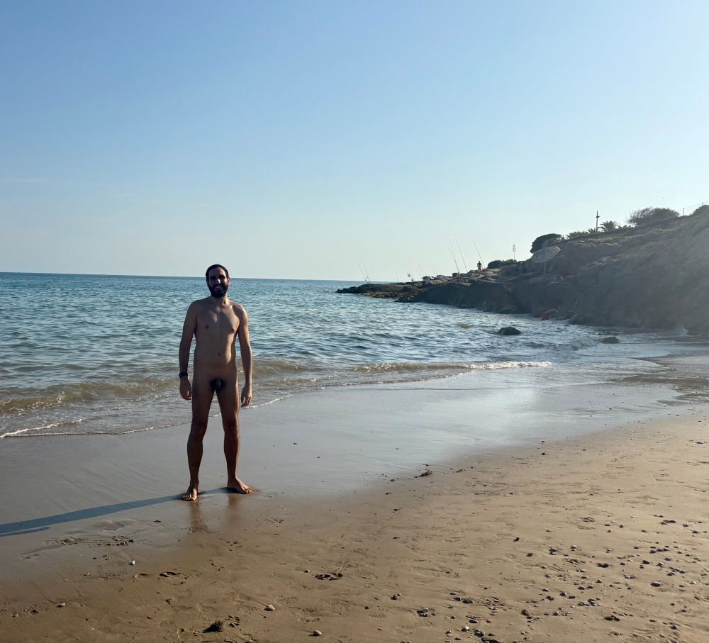 A naked person standing at the beach, with the sea and rocky cliffs in the background.