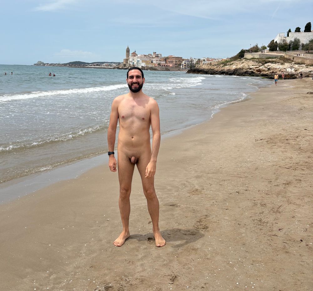 A nude, smiling person standing on the beach, with a town visible in the background.