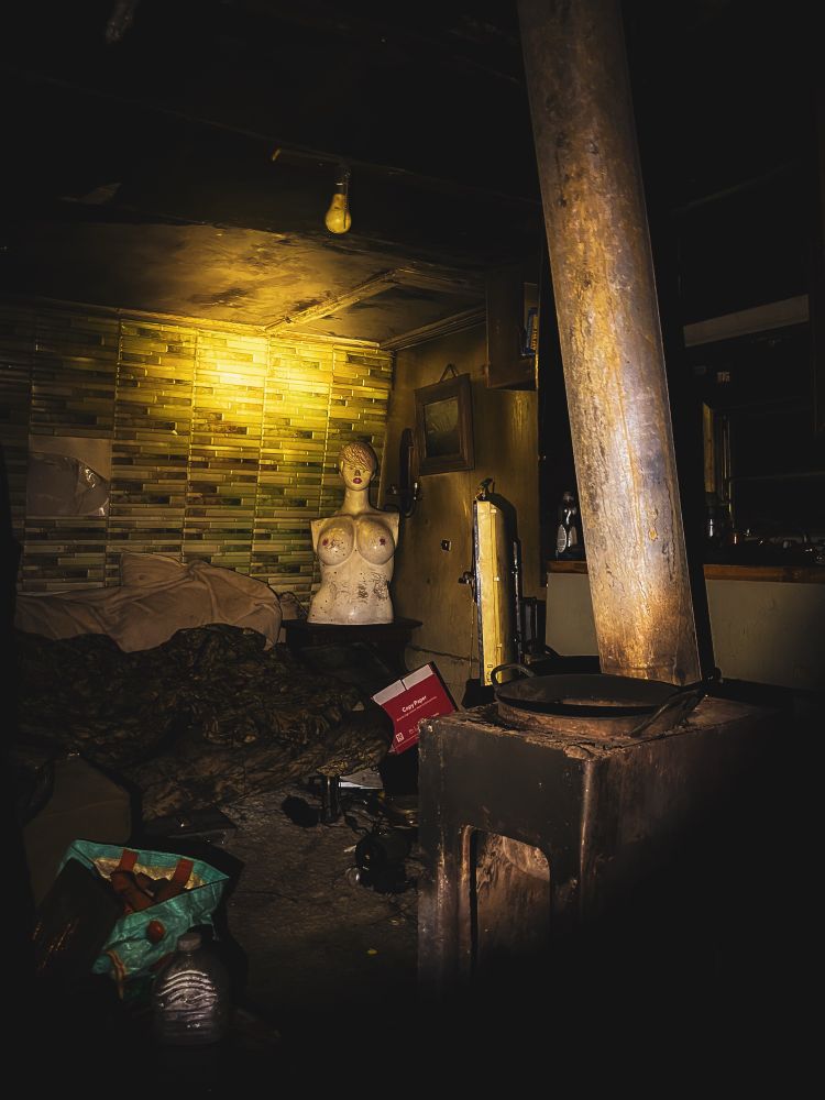 Dark photo of the inside of an old RV. There is a stove and lots of litter on the floor. The highlight is a very busty mannequin in the back corner.