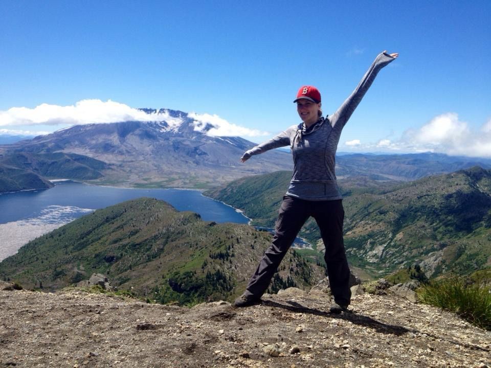 Woman standing and pointing in front of an overlook of Mount St Helens volcano on a blue sky day.
