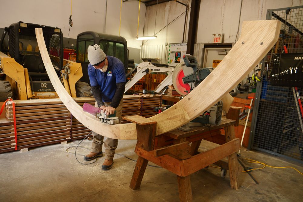 Inside a large workshop with landscaping vehicles parked in the background, a man wearing a dust mask uses a power sander on a long arching wooden beam.  
