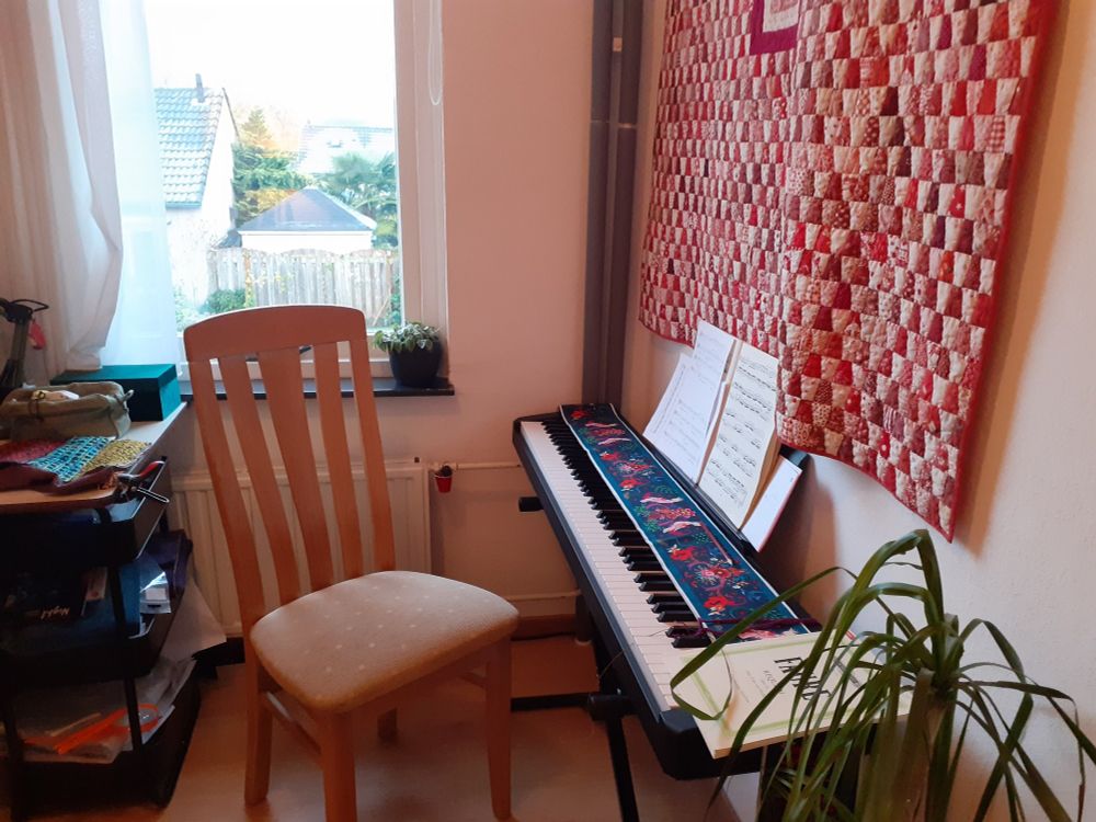 Digital piano alongside a wall, the Requiem of Faure (for choir) and some other sheet music on it. A kitchen chair, a window with a white curtain and a little plant. The wall hanging is a red-white quilt.