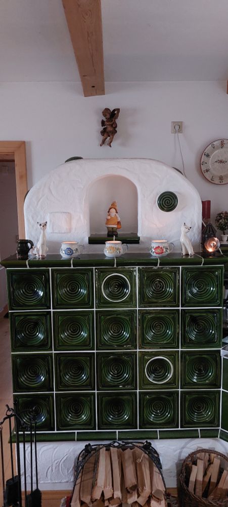 Kachelofen im klassischen Grün mit weißen Sockel . Darauf stehend 3 Tassen in verschiedenen Farben ( blau, gelb, rot mit Blumenmuster) 2Keramikkatzen, 1 alte Grubenlampe eine Keramikfrau in der Mitte des aufgesetzen Gugl und eine kleine Vase. Darüber schwebend, über dem Kachelofen, ein Engerl aus Holz.