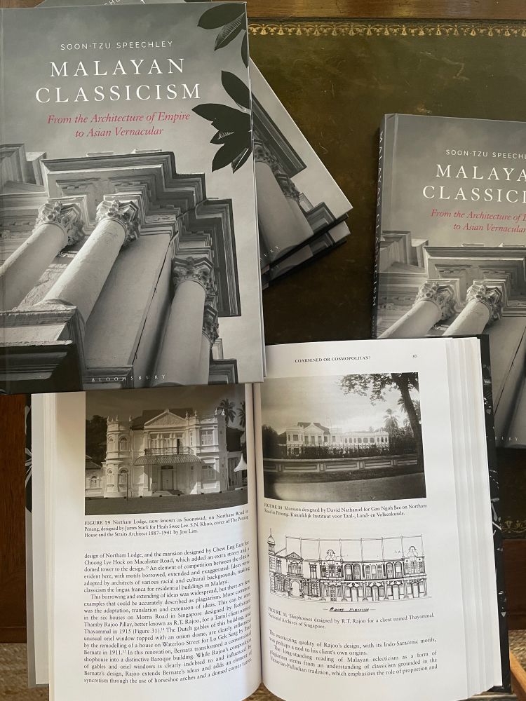 Copies of the book Malayan Classicism on an antique desk, with one copy open to a two-page spread featuring black and white photographs and a building plan.