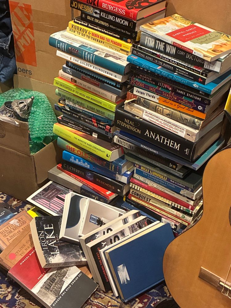 To Be Read books, two stacks and a loose pile on the floor in front, maybe 50 total, and the edge of an acoustic guitar.