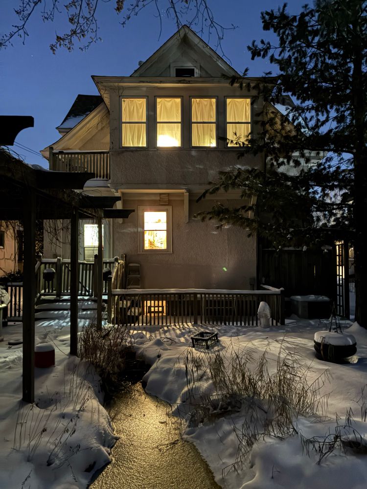 Two story house at night, lit windows reflecting on snow and frozen pond.