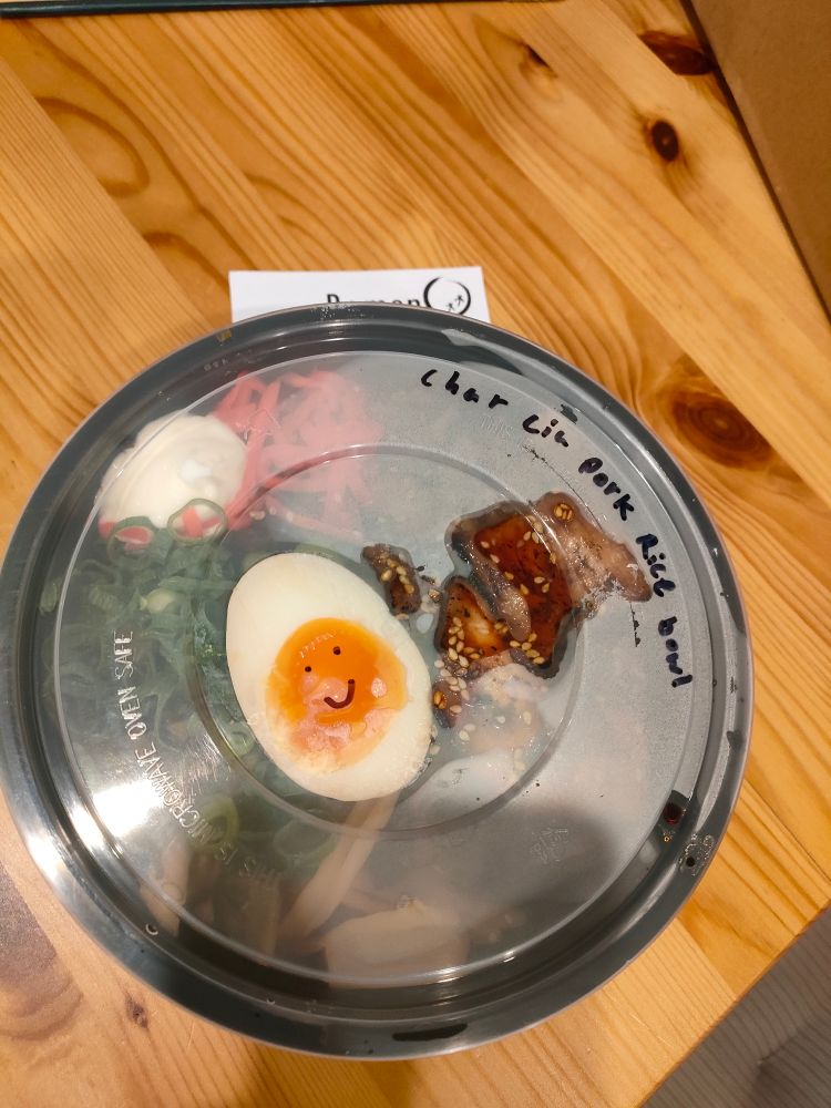 A large plastic takeaway bowl with 'Char ciu pork rice bowl' written on the transparent lid. A smiley is drawn on the lid above the yolk of half a boiled egg.