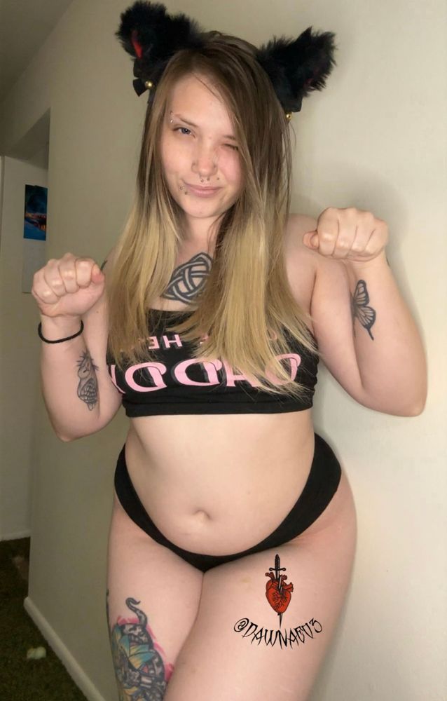 Model is posing with her arms up in a shape of a fist like a cat! She’s winking at you.
She’s wearing something cute that consist of two different pieces that are both black with pink lettering.
She is also wearing blacking cat ears that have red lining with little gold bells on the bottom!