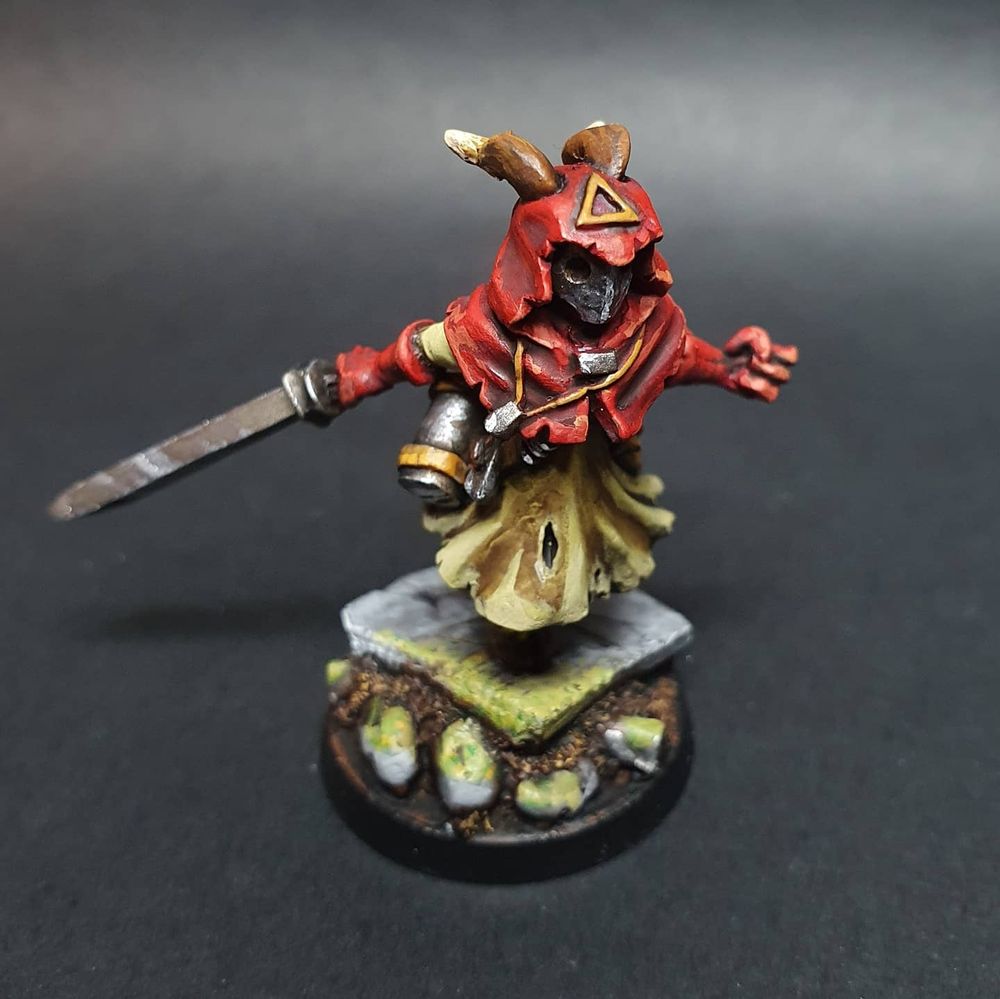 A red hooded fantasy miniature with horns and a masked face. Theybare holding a sword and seem to be leaping forward.