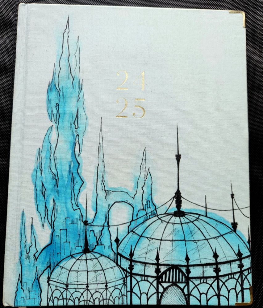 A picture of a blue planner notebook with art deco buildings and a crystal tower drawn on it in ink and watercolor. 24-25 is printed on the cover.