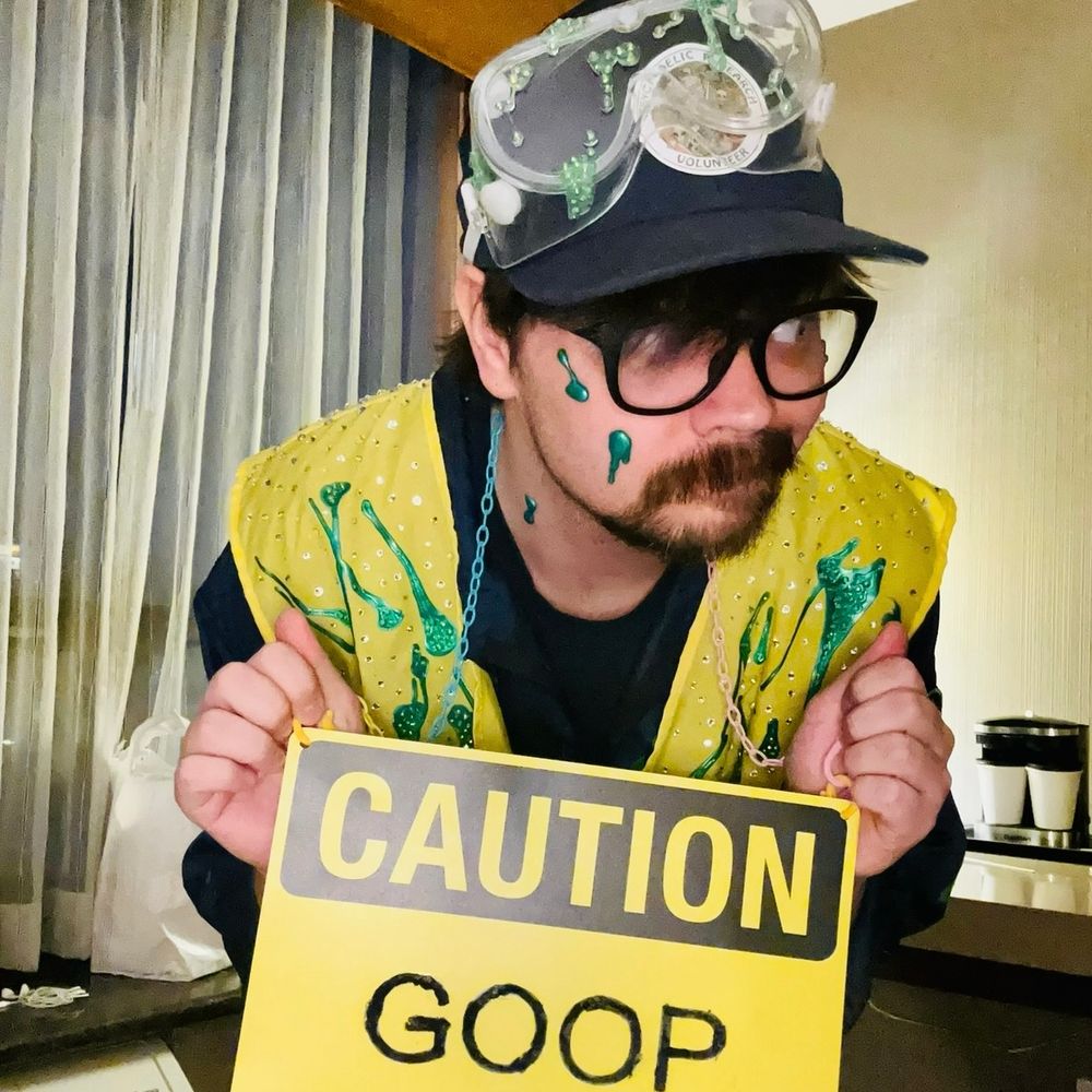 Will’s look for Countdown 2024. Alien clean up crew look with overalls and saftey vest covered in sparkly green goop holding a sign that reads “Caution: Goop”