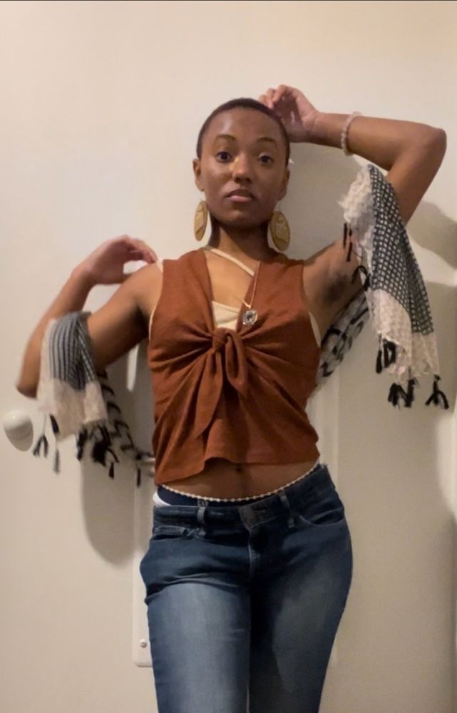 Dia, a brown skinned Black person is posing in low rise jeans, a brown sleeveless sweater, & a scarf draped across their arms 