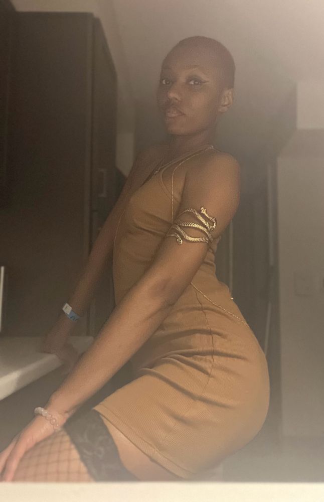 Dia, posing from thigh-up, wearing a fishnet stocking, a skin toned brown dress, and gold toned jewelry 